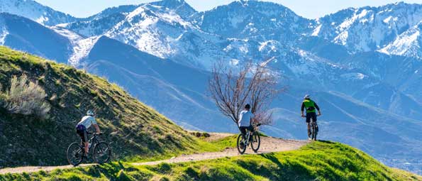 Three mountain bikers ride a trail overlooking the snow-capped Wasatch mountains