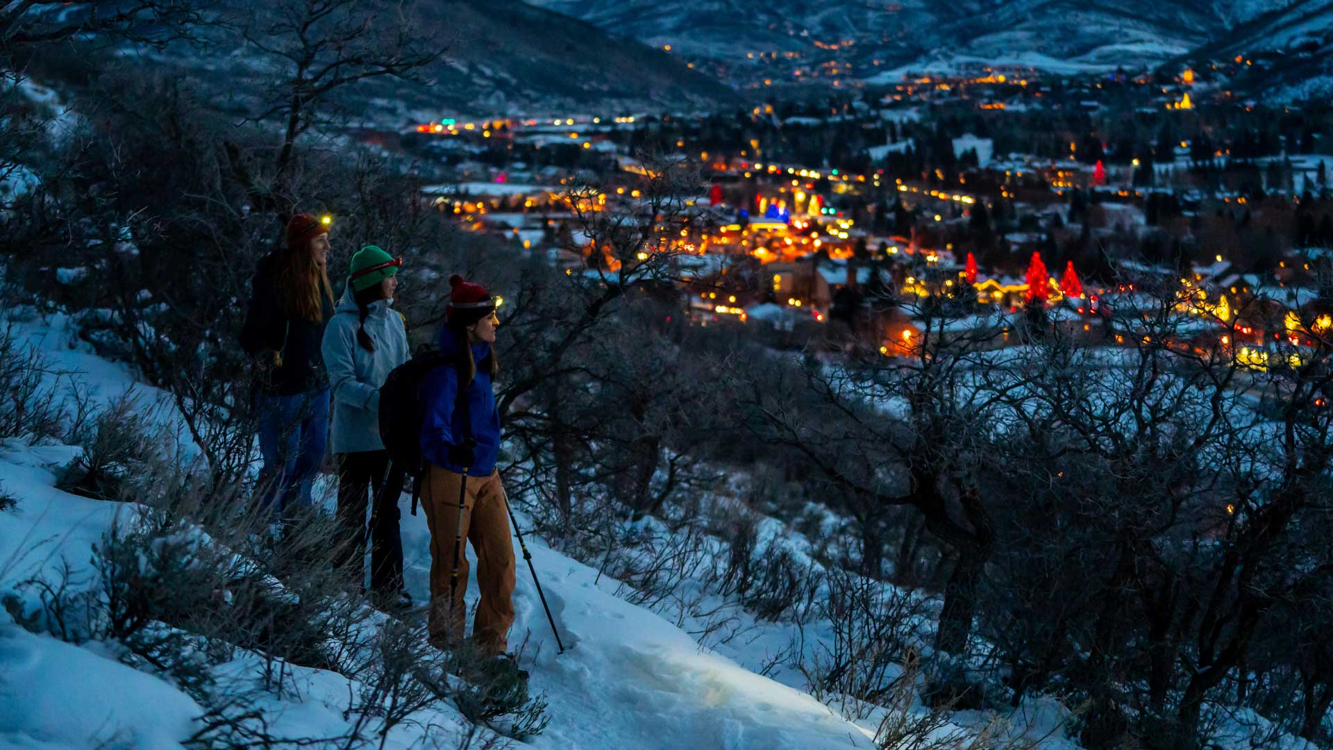 Three snowshoers overlook the lit-up town of Park City, UT at twilight