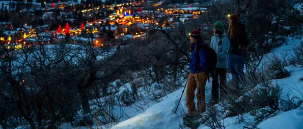 Three snowshoers overlook the lit-up town of Park City, UT at twilight