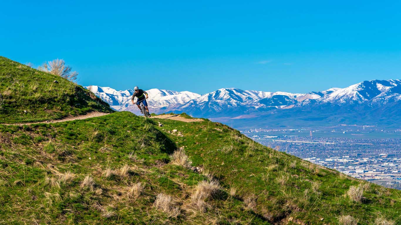 A mountain biker rides a trail surrounded by green grass and snow capped mountains in Salt Lake City, UT